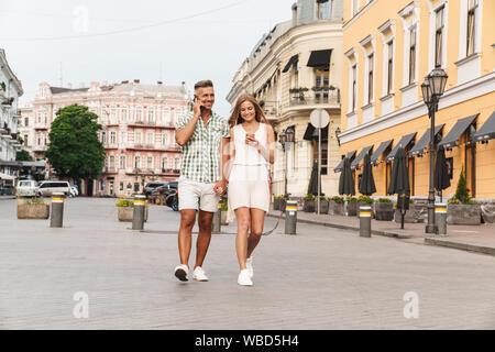 Image of beautiful happy couple in summer clothes holding hands together and using smartphones while walking through city street Stock Photo