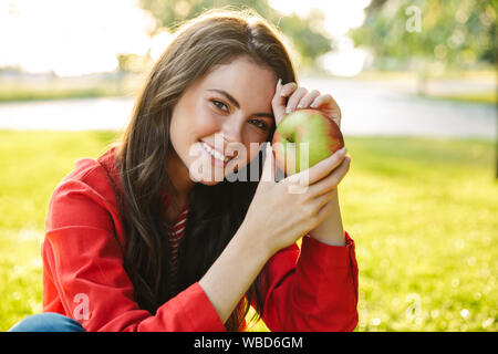 Image of nice girl student wearing red jacket smiling and holding apple while sitting in green park Stock Photo