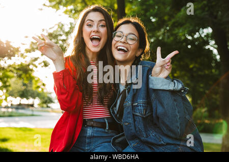 Image of two laughing girls students gesturing peace sing and hugging while sitting on railing in green park Stock Photo