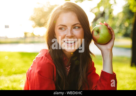 Image of lovely girl student wearing red jacket smiling and holding apple while sitting in green park Stock Photo