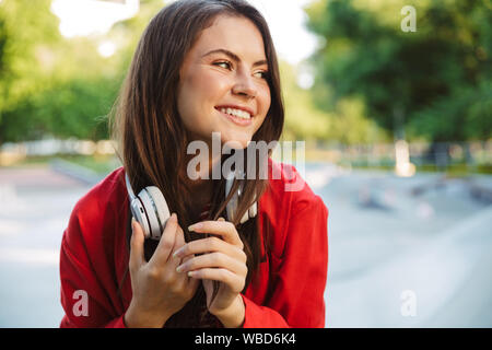 Image closeup of pleased girl student wearing red jacket poising with headphones over neck on sports ground Stock Photo