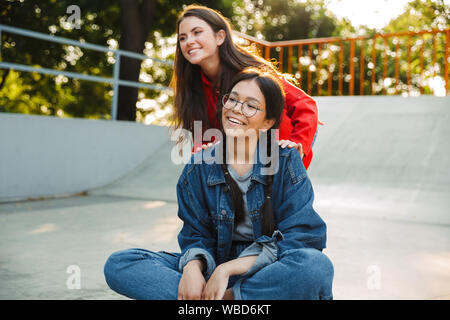 Image of two pretty girls dressed in denim wear laughing and riding skateboard together in skate park Stock Photo