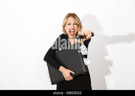 Portrait of an attractive blonde woman dressed in black dress standing isolated over white background, holding a gun, shooting while holding briefcase Stock Photo