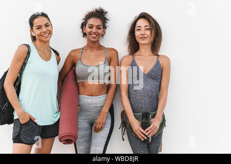 Photo of beautiful multiethnic sportswomen in tracksuits smiling and standing over white wall outdoors Stock Photo