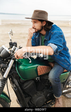 Handsome young man wearing casual outfit sitting on a motocycle at the beach, wearing a hat Stock Photo