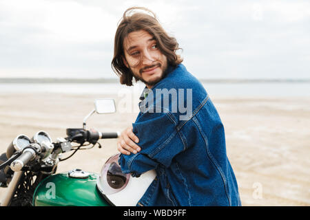 Image of a handsome positive optimistic happy man biker on his bike outdoors on a beach looking away. Stock Photo