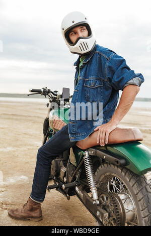 Handsome young man wearing casual outfit sitting on a motocycle at the beach, wearing a helmet Stock Photo