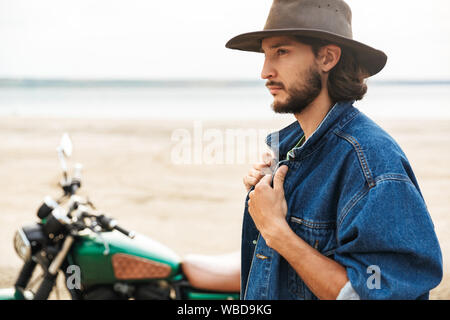 Close up of a handsome young man wearing casual outfit standing at motocycle at the beach Stock Photo