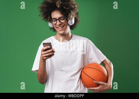 Portrait of cheerful man with afro hairstyle playing using cellphone and headphones while holding basketball isolated over green background Stock Photo
