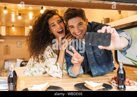 Portrait of happy couple man and woman gesturing peace sign while taking selfie photo on smartphone in cozy kitchen at trailer indoors Stock Photo