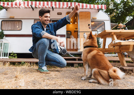 Portrait of unshaven handsome man wearing denim clothes playing with red dog near house on wheels outdoors Stock Photo