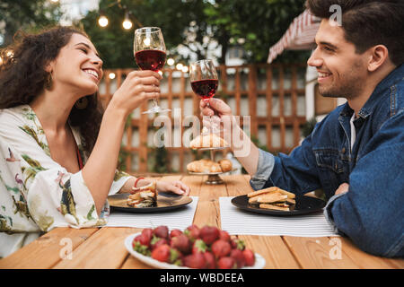 Portrait of smiling couple man and woman drinking red wine while eating sandwiches together at wooden table outdoors Stock Photo