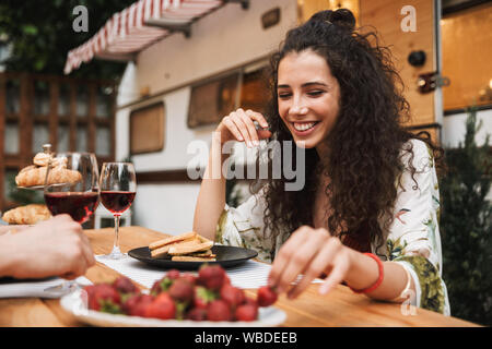 Portrait of cute couple man and woman drinking red wine while eating strawberry together at wooden table outdoors Stock Photo