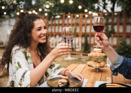 Portrait of pleased couple man and woman drinking red wine while eating sandwiches together at wooden table outdoors Stock Photo