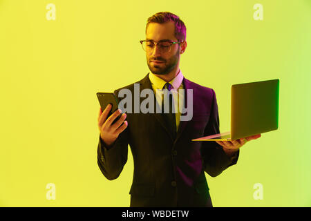 Image of a handsome young business man posing isolated over light green background wall with led neon lights using laptop computer using mobile phone. Stock Photo