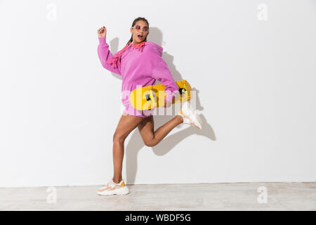 Image of amazing young african woman running isolated over white wall background in bright pink sweatshirt holding skateboard looking away. Stock Photo