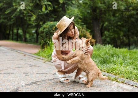 Portrait of nice woman wearing dress stroking dog and making kiss face while squatting in summer park Stock Photo