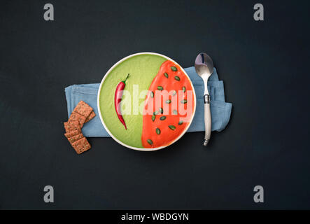 Cream soup bowl with crackers on black background. Red and green soup plate. Tomato and spinach cream soup. Flat lay of healthy food. Vegetarian meal Stock Photo