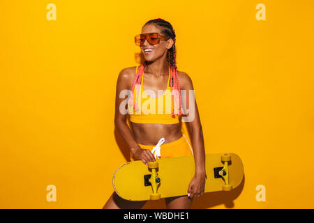 Image of nice african american woman wearing sunglasses holding skateboard and laughing isolated over yellow background Stock Photo