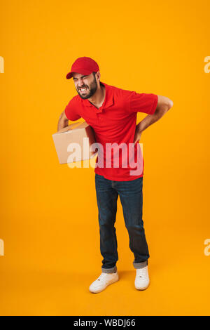 Full length portrait of upset delivery man in red uniform touching his painful back while holding packaging box isolated over yellow background Stock Photo