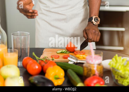 Closeup cropped shot of african man preparing vegetable salad, holding knife, cutting tomatoes on chopping board in the kitchen at home. Vegetarian, h Stock Photo
