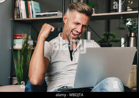 Photo of excited adult man in casual clothing smiling and celebrating while using laptop in apartment Stock Photo