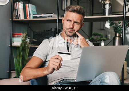 Photo of thoughtful adult man in casual clothing holding credit card and laptop while sitting on armchair in apartment Stock Photo