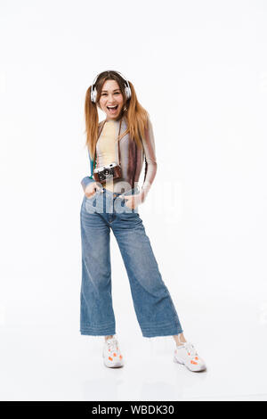 Full length photo of pretty young girl wearing headphones laughing and carrying retro camera isolated over white background Stock Photo