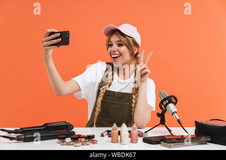 Image of teen girl taking selfie on smartphone and recording blog broadcast about new cosmetic products isolated over orange wall in studio Stock Photo