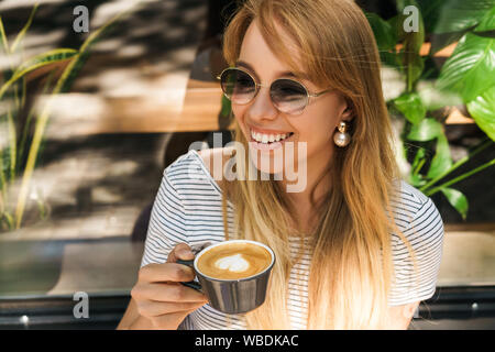 Portrait of fashionable young woman wearing sunglasses smiling and holding coffee cup while sitting in street cafe outdoors Stock Photo