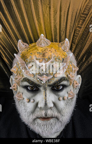 Head with thorns or warts, face covered with glitters, close up. Demon with golden collar, black background. Alien, demon, sorcerer makeup. Senior man Stock Photo