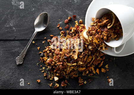 Heap of dried fruit tea infusion with oranges and strawberries mixed with tea leaves and assorted herbs in tea cup and spoon on black wooden table bac Stock Photo
