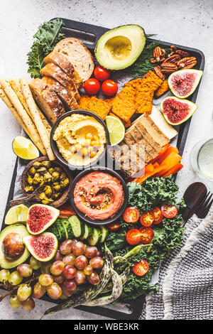 Vegan appetizer platter. Hummus, tofu, vegetables, fruits and bread on a black tray, white background. Stock Photo
