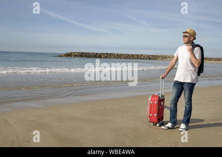 Attractive male traveler in a hat and with a red suitcase stands on the seashore, taking off his jacket, copyspace Stock Photo