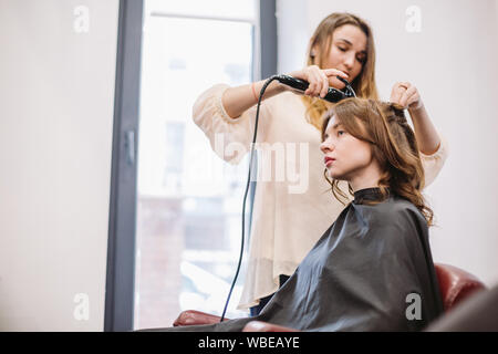 beauty, hairstyle concept, happy young woman and hairdresser with hair iron making hairdo at hair salon. Woman Having Hair Stylized Hairdresser Stock Photo