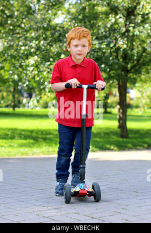 Child learn to ride scooter in a city park on sunny summer day. Cheerful little boy with ginger hair posing with a scooter. Active leisure and outdoor Stock Photo