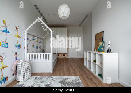 Contemporary children room with white walls with colorful images and a parquet with carpet on the floor. There is a bed, shelves with books and toys, Stock Photo
