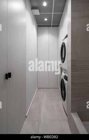 Contemporary laundry room with textured beige tiled walls and luminous lamps. There are light lockers, two washing machines which stand on each other. Stock Photo
