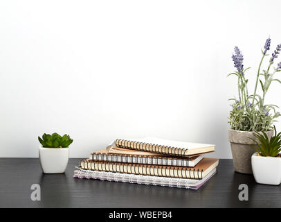 stack of spiral notebooks with white pages and ceramic pots with plants on a black table, white wall, minimalism in Scandinavian style Stock Photo