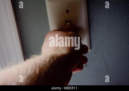Hand flipping light switch against a grey blue wall with faint light Stock Photo