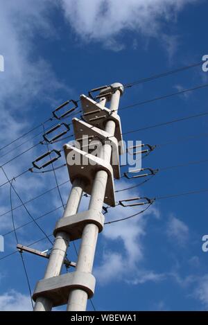 An concrete electrical pylon stands tall against the blue sky. Ceramic insulators can be seen holding the wires on both sides of the structure. Stock Photo