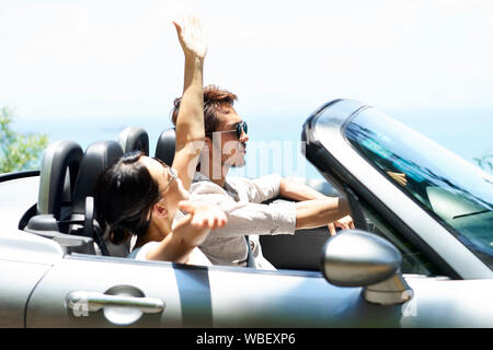 young asian couple riding in a convertible car on a seaside road, side view