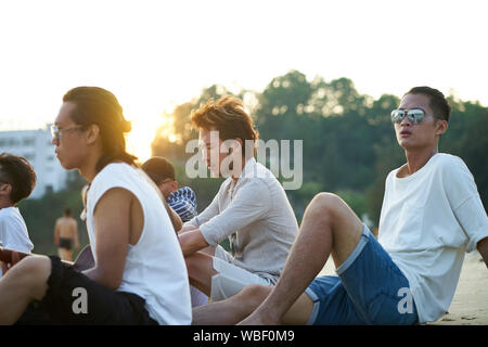 group of five young asian men sitting on sand beach at dusk Stock Photo