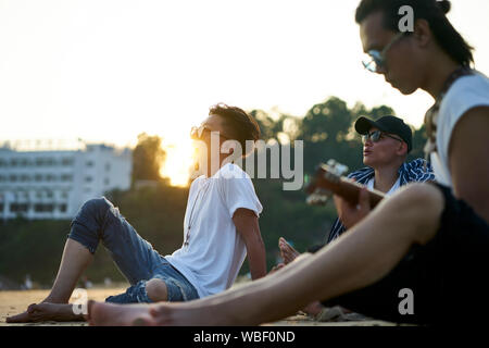 three young asian adult men sitting on sand beach playing guitar at dusk Stock Photo