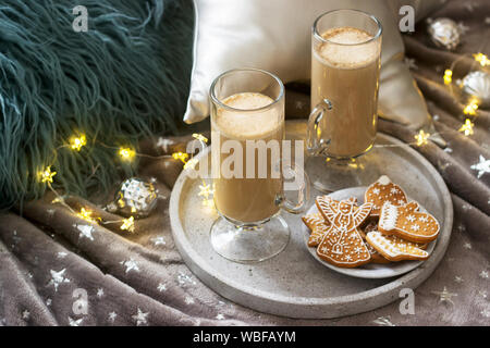 Homemade latte in glass cups, served with gingerbread on the background of plaid, pillows and garlands. Stock Photo
