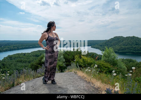 Fashionable Woman of 30-40 years old outdoors standing tall on a hill with a river running through the woods under a blue sky in the background Stock Photo