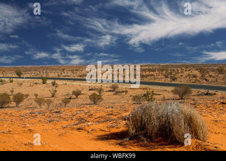 Road stretching across vast red Australian outback plains with scattering of low dry vegetation under blue sky Stock Photo