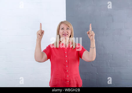 For comparison mature woman points her fingers up while standing against a half of a gray and white brick wall. Stock Photo