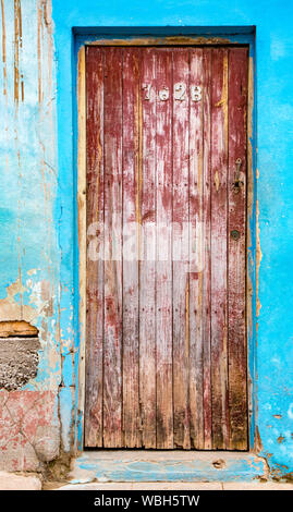 Trinidaad, Cuba Nov 26, 2017 - Faded red door on blue brick wall with broken plaster Stock Photo