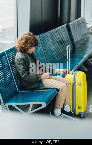 side view of boy holding passport and sitting on blue seat near yellow suitcase in waiting hall in airport Stock Photo